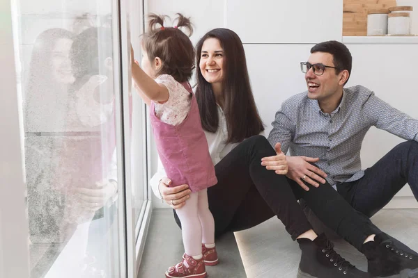 A happy family is sitting by the window. The baby is looking out the window. Parents look at their daughter in love with eyes. Joyful dad and mom are watching the baby