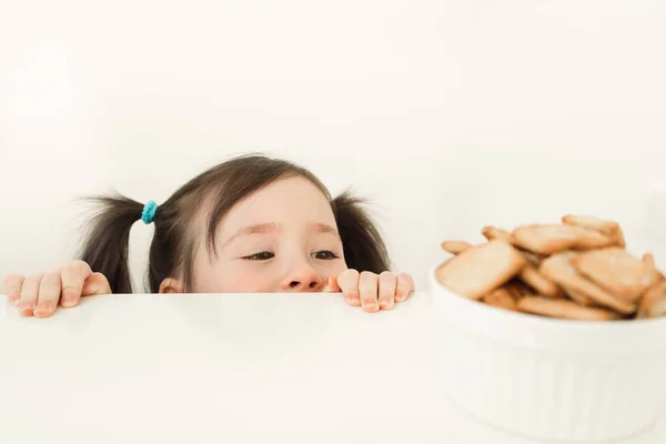 A child spying on cookies. Baby wants to steal sweets. Girl peeks out from the table — Stock Photo, Image