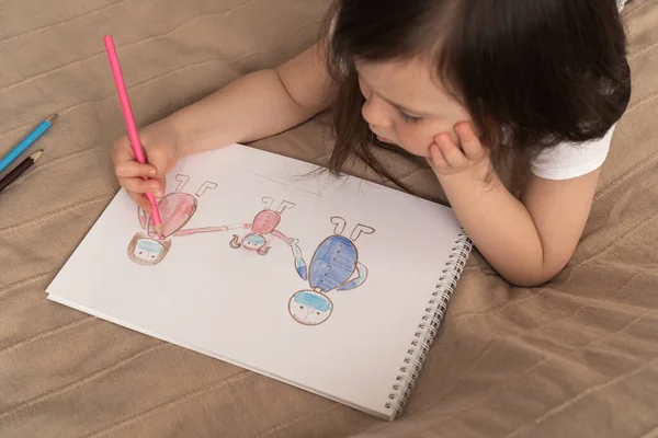 Cute girl draws a pencil drawing. A child learns to draw people. The child lies on the bed and draws with a pink pencil.