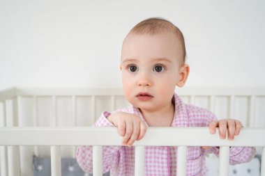 Güzel pembe pijamalı bebek. Çocuk uyandı ve çok şaşırmış görünüyordu. Bir çocuk beyaz beşiğinde duruyor.