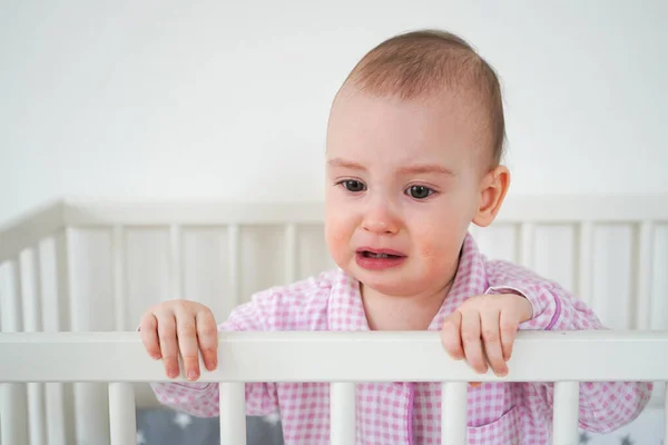 A small child in pink pajamas is standing in the crib. The kid woke up and waits for his parents to pull him out of the crib. Baby starts to cry.