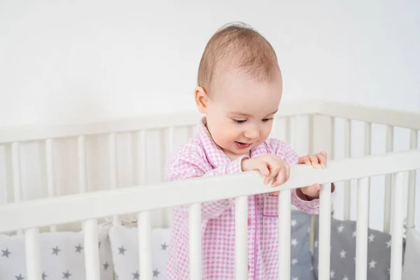 A small child in pink pajamas stands in the crib. The kid woke up and waits for his parents to get him out of the crib