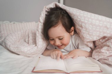 Çocuk yatmadan önce yatağında bir kitap okur. Kız yorganın altına saklandı ve okudu. Pembe bir battaniyede saklanan bir bebek.