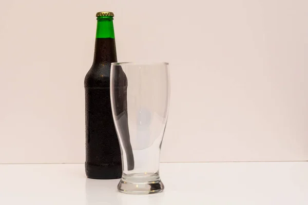 Vidro transparente vazio e uma garrafa verde fechada com cerveja escura em um fundo claro — Fotografia de Stock