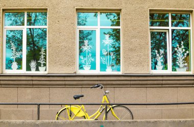 Binanın ön cephesinde, pencereleri taklit çiçeklerle süslenmiş sarı bisiklet ve yansıması mavi gökyüzü..