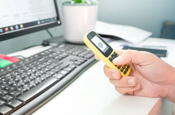 Beyaz masaüstünde oturan, eski moda basma tuşlu cep telefonuyla yakın plan erkek eli. Evde kalmak, uzaktan çalışmak, ev ofisinden uzak durmak ve fırfırlı iş kavramı.