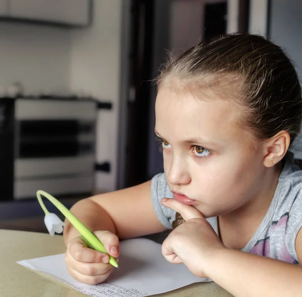 Porträtt av en seriös grundskoleflicka som skriver ett brev på ett papper med pennan i handen sittande vid bordet. Önskelista, tänkande över idé, koncentration, komponera ett dikt koncept — Stockfoto