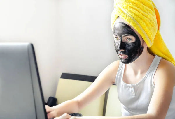 집에 있는 랩탑 컴퓨터를 사용하는 10 대 소녀가 검은 얼굴의 진흙 마스크와 노란 수건을 머리에 쓰고 있습니다. 집에 머물고, 격리되고, 거리 학습을 하고, 온라인 교육을 받고, 기술을 배우고, 현대 생활 방식을 유지하고, — 스톡 사진