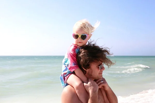 Toddler dziewczynka siedzi na ramionach ojca na plaży przez th — Zdjęcie stockowe