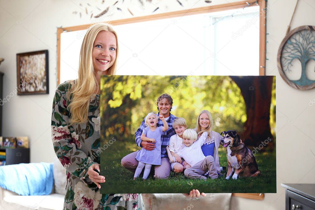 A happy young blonde woman is holding a large wall canvas portrait of her family with young children and a pet dog.