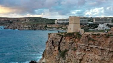 Ghajn Tuffieha Kulesi ve sahil havadan görünüyor. Gün batımı, mavi gökyüzü, yeşil tepe, denizde dalgalar. Kış. Kamera hareket çemberi. Malta