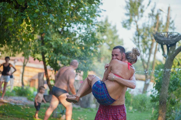 Feliz pareja joven se divierte en un día caluroso de verano jugando con el aspersor de manguera de agua en el jardín. Two Young People in Love Got Wet Brokingly Fighting with Hose. En cámara lenta. El hombre salva a la chica de th — Foto de Stock