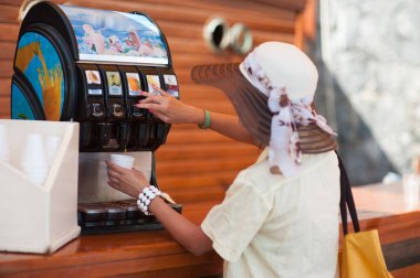 Kız makineden bir bardağa su dolduruyor, kumsalda susuzluk çekiyor.
