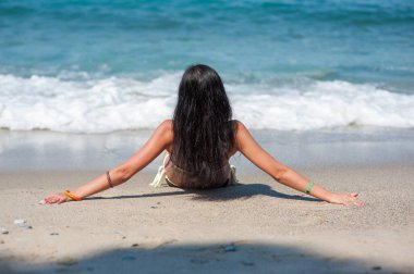 Esmer genç kız kumsalda güneşleniyor, dalgalar bacaklara vuruyor.