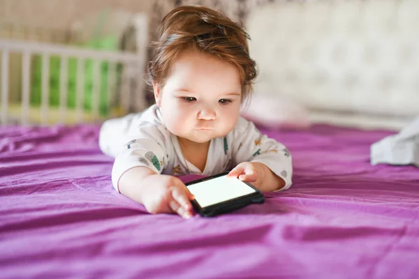 Детская зависимость от телефонов. излучение от телефона ребенку. Маленький мальчик 0-1 лет со смартфоном в руках с энтузиазмом смотрит на экран. Детская гаджетная зависимость — стоковое фото