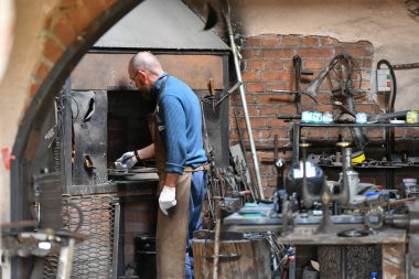 Deneyimli bir demirci, açık ateşle çalışır. Demirci ocaktaki alevi havai fişekle söndürür, atölyede kızgın demir döver.