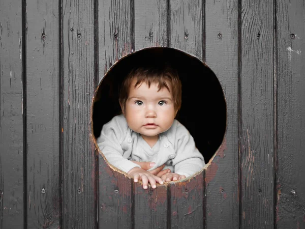 Bambino guarda attraverso un foro rotondo. sfondo in legno scuro. sorride alla telecamera. posto per iscrizioni. ritratto di bambino felice, il bambino sta guardando fuori dalla finestra di legno rotonda e sorridente — Foto Stock