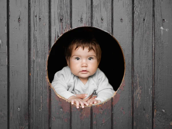 Kind kijkt uit een rond houten raam en glimlacht. De jongen klom uit het ronde raam en glimlacht vrij recht in het frame. Vrolijk, vrolijk, speels, nieuwsgierig jongetje. kind 0-1 jaar — Stockfoto