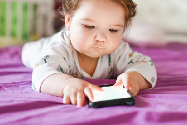 Вред от мобильного устройства ребенку. Малыш Тоддлер лежит, глядя на телефон. Концепция зависимости от гаджетов — стоковое фото