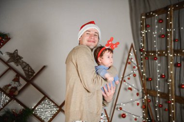 Baba ve oğul Noel ağacında kucaklaşıyorlar. Mutlu bir çocuk ve babası çerçeveye bakıyorlar. Baba oğluyla Noel'de kucaklaşmak. ve eğlenin. kafasına boynuzları. Yeni Yıl şapkası.
