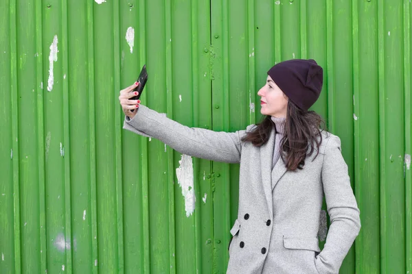 Meisje neemt een selfie op een groene achtergrond. een hoed en jas dragen. Een plek om te schrijven. gekleed in een koude pagode. kleren en stijl. natuurlijke emoties — Stockfoto