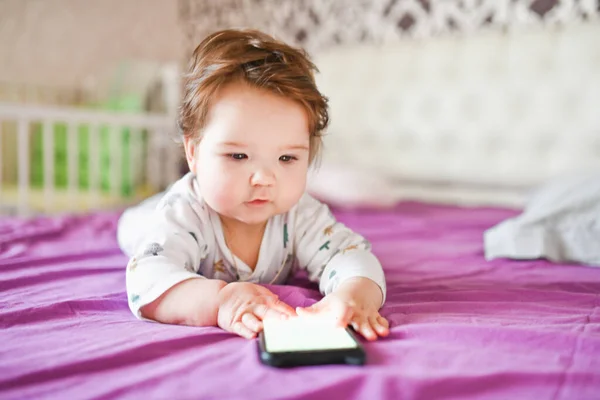 Gyermek és mobil eszköz. Egy kislány az ágyban, aki okostelefont néz. — Stock Fotó
