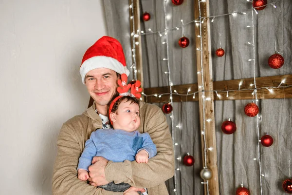 Noel geçmişinde çocuklu bir baba. Küçük bir çocuğun kafasında kırmızı boynuzlar. Yeni yıl. Noel kutlaması. Güzel bir aile Noel ağacının yanında oturur. Süveter giyen yakışıklı adam. Küçük çocuk. — Stok fotoğraf