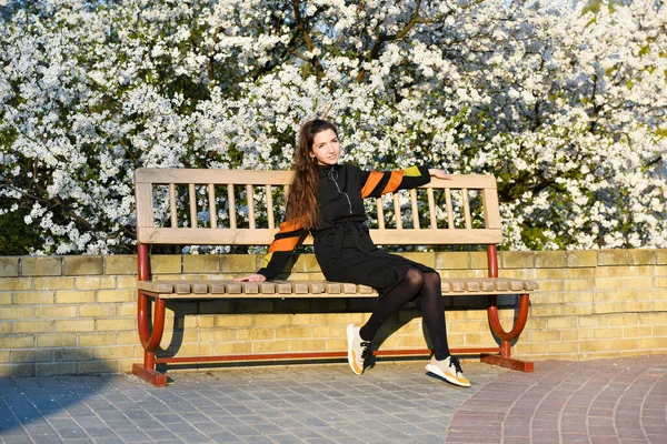 Meisje op het glimlachen gezicht zit op de Bank, sakura op achtergrond, intreepupil. Meisje ontspannen terwijl wandeling in park in de buurt van kersenbloesem. Mode accessoires concept. Schattig kind met rugzak genieten van zonnige lente — Stockfoto