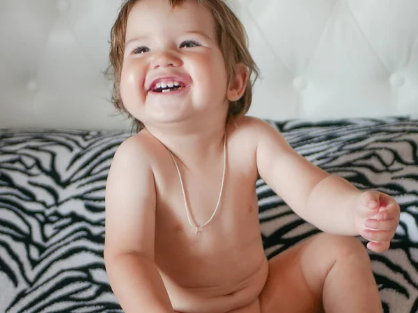 De jongen lacht vrolijk op het bed. Met een naakte romp. Vrolijk kind. Het kind zit op het bed en lacht, plezier kijkend naar de top. — Stockfoto