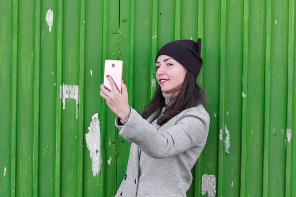 Chica toma una selfie en un fondo verde. con sombrero y abrigo. lugar para escribir. vestida con una pagoda fría. ropa y estilo. emociones naturales — Foto de Stock