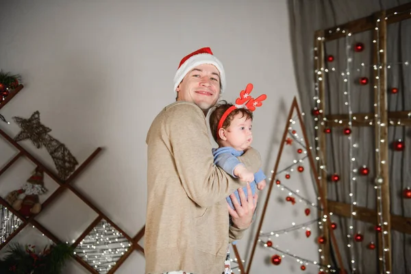 Baba ve oğul Noel ağacında kucaklaşıyorlar. Mutlu bir çocuk ve babası çerçeveye bakıyorlar. Baba oğluyla Noel'de kucaklaşmak. ve eğlenin. kafasına boynuzları. Yeni Yıl şapkası. — Stok fotoğraf