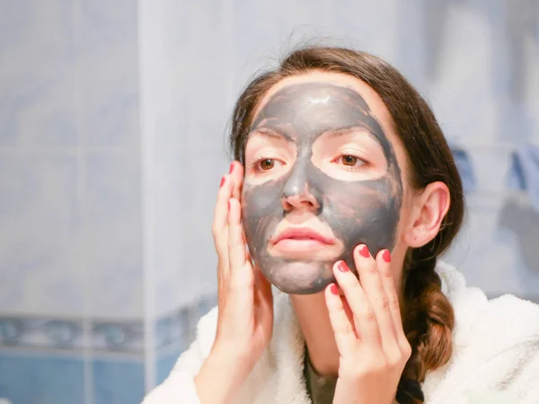 Cosmetic black mask. Woman brings beauty to her face, gets rid of black dots, using a black mask. A female in the bathroom looks in the mirror — Stock Photo, Image