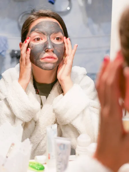 A menina na máscara facial olha no espelho descontente. Máscara preta cosmética. Mulher traz beleza para o rosto, se livra de pontos pretos, usando uma máscara preta. Uma fêmea no banheiro olha no espelho — Fotografia de Stock