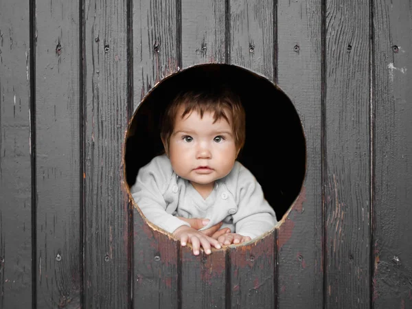 Ребенок смотрит через круглую дыру. темный деревянный фон. улыбается в камеру. место для надписей. Портрет счастливого маленького мальчика, малыш смотрит в круглое деревянное окно и улыбается — стоковое фото