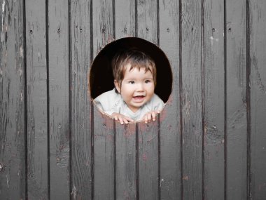 Çocuk yuvarlak bir delikten bakıyor. Koyu ahşap arka plan. Kameraya gülümsüyor. Yazıtlar için yer. Küçük mutlu çocuğun portresi, çocuk yuvarlak ahşap pencereden dışarı bakıyor ve gülümsüyor.
