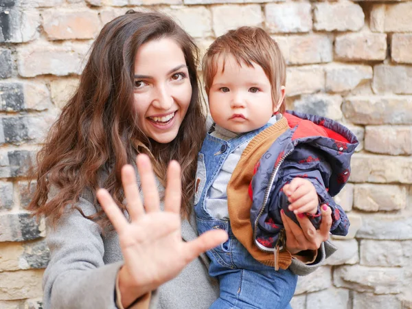 Tuğla arka planda annesi olan bir bebek. Kalın giysiler içinde kameraya bakıyorum. Mutlu ve genç bir anne tuğla zeminde oturuyor ve bebeğini kucağında tutuyor. Aşk ve aile kavramı. Genç anne elinde tutuyordu. — Stok fotoğraf