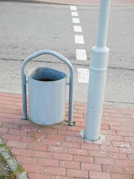 Urne aus Metall neben der Straße. Ein metallener Mülleimer steht neben einer Säule auf dem Bürgersteig. Schmieden von Mülltonnen steht neben einer Bank auf dem Bürgersteig — Stockfoto