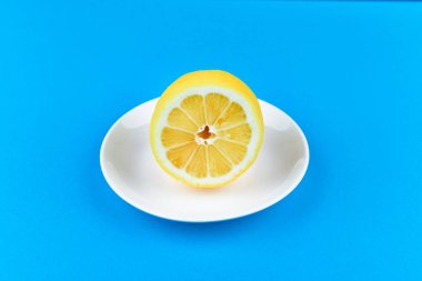 Mavi arka planda limon. Beyaz bir tabakta yatıyor. Yarısı kesilmiş limon. Yukarıdan bak. Kayıt için bir yer. Mavi arka planda limon dilimleri. Yazın, serin portakal dilimleri. Limon, meyve, ferahlatıcı, sarı