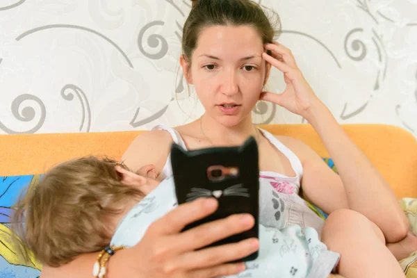 Mom feeds the baby and looks at the phone. A young mother with a small child in her arms holds a smartphone and looks at the screen, uses electronic devices, the child is sad. the affection of young m — Stock Photo, Image