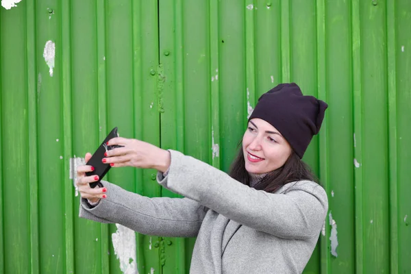 Meisje neemt een selfie op een groene achtergrond. een hoed en jas dragen. Een plek om te schrijven. gekleed in een koude pagode. kleren en stijl. natuurlijke emoties — Stockfoto