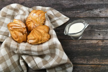 Koyu ahşap arka planda sütlü Choux keki. Kareli havlunun üzerinde bir sepetin içinde. Yukarıdan bak. Süzme peynirli şık. Hasır kasede küçük muhallebi kekleri ahşap arka planda