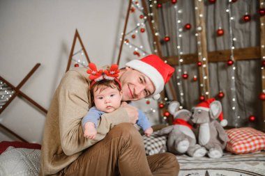 Baba ve oğul Noel ağacında kucaklaşıyorlar. Mutlu bir çocuk ve babası çerçeveye bakıyorlar. Baba oğluyla Noel'de kucaklaşmak. ve eğlenin. kafasına boynuzları. Yeni Yıl şapkası.
