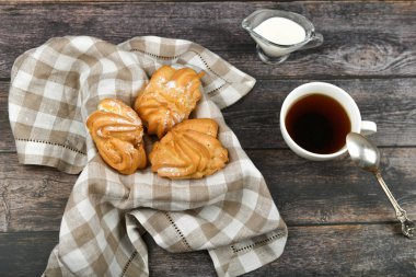 Ahşap zemin üzerinde kahve olan Choux keki. Kareli havlunun üzerinde bir sepetin içinde. Yukarıdan bak. Süzme peynirli şık. Hasır kasede küçük muhallebi kekleri ahşap arka planda