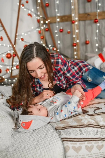 Yılbaşı atmosferinde çocuklu genç bir anne. Noel ailesi, anne ve bebek Noel ağacının yanında gülümsüyor. Oturma odası Noel ağacı ve hediye kutusuyla süslenmiş. — Stok fotoğraf