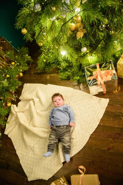 Dziecko w pobliżu choinki widok z góry. Portret zbliżeniowy uśmiechniętego dziecka w świątecznym wnętrzu w pobliżu świątecznej choinki. Widok z góry szczęśliwy dzieciak leżący na białej drewnianej podłodze w pobliżu prezenty unde — Zdjęcie stockowe