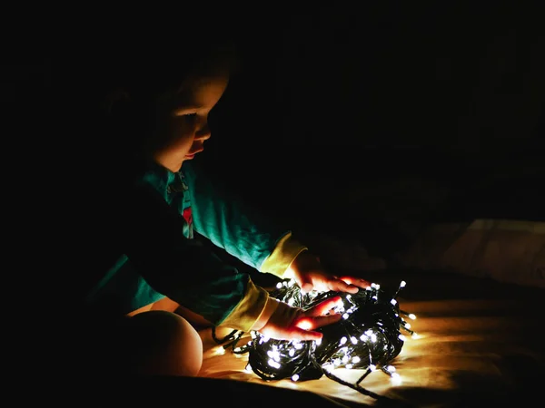 Niño y huerta en la oscuridad. el niño pequeño sostiene luces brillantes de christmas interiores cerrar fondo oscuro. Retrato de un niño feliz sonriendo a un huerto festivo en vísperas de las vacaciones de las manos. El —  Fotos de Stock