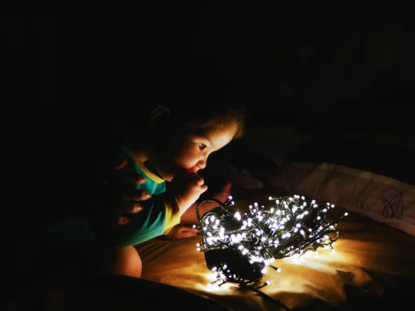 child and garland in the dark. little child holds shiny christmas lights indoors close dark background. Portrait of a happy kid boy smiling to a festive garland on the eve of the holiday of hands. The