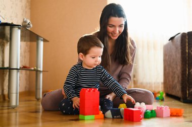 Anne ve bebek yerde oynuyorlar. Güzel bebek oyuncaklarla oynuyor. Mutlu anne içeride.