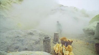 JAVA, INDONESIA - JULY 7, 2019: Indonesian man without respirator working on loading toxic sulphur into baskets surrounded by poisonous fumes of Ijen volcano 