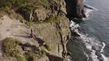 Okyanusun yukarısındaki yüksek kayalıklarda duran güzel elbiseli çingene kadının hava panoramik görüntüsü.